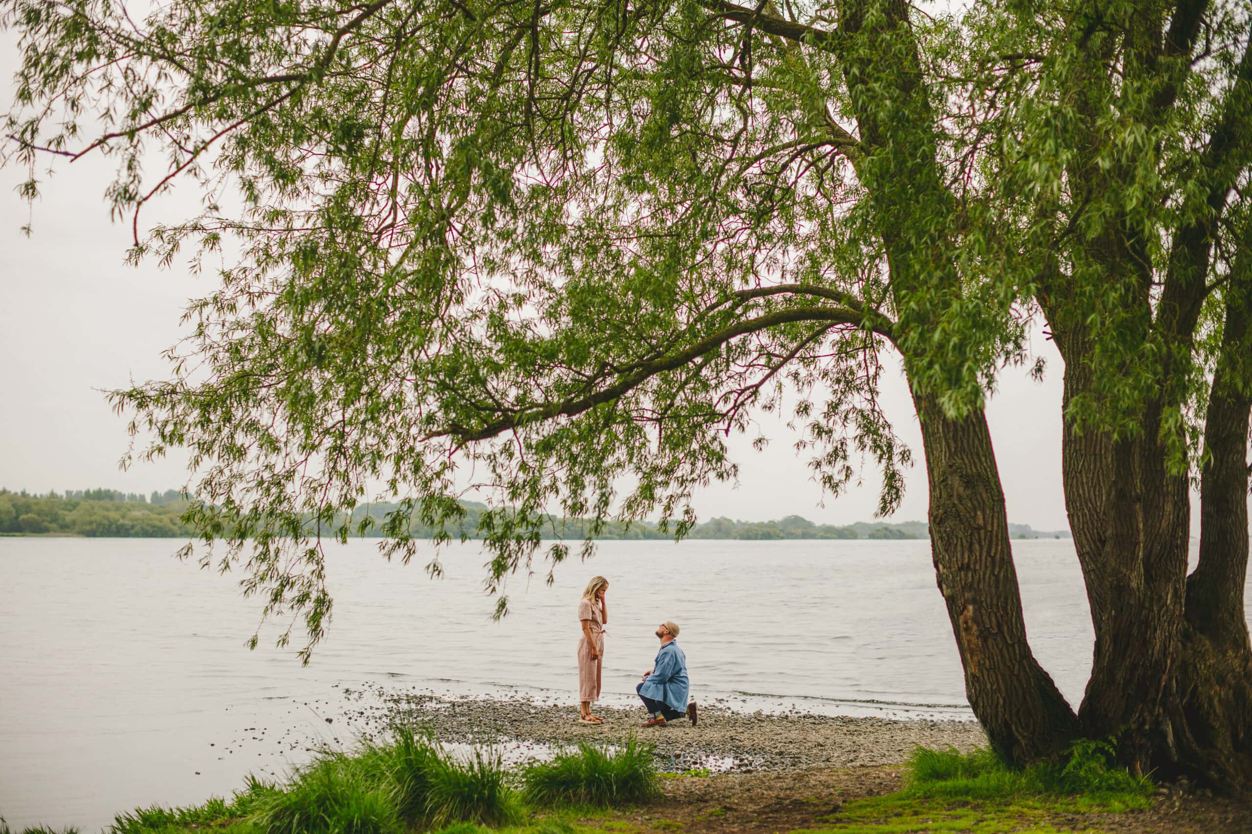 Couple framed by tree standing in front of a lough while man is down on one knee proposing to the girl