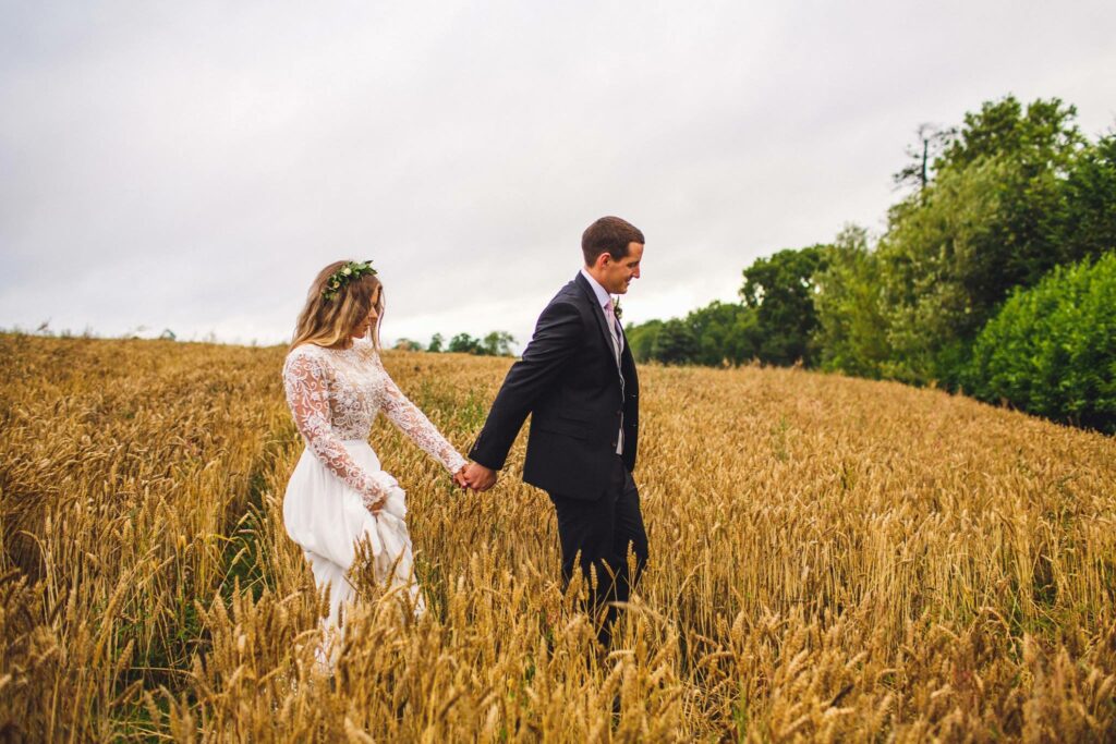 A stunning wedding photo captures a beaming bride and groom walking hand in hand through the lush golden wheat field at Gracehall