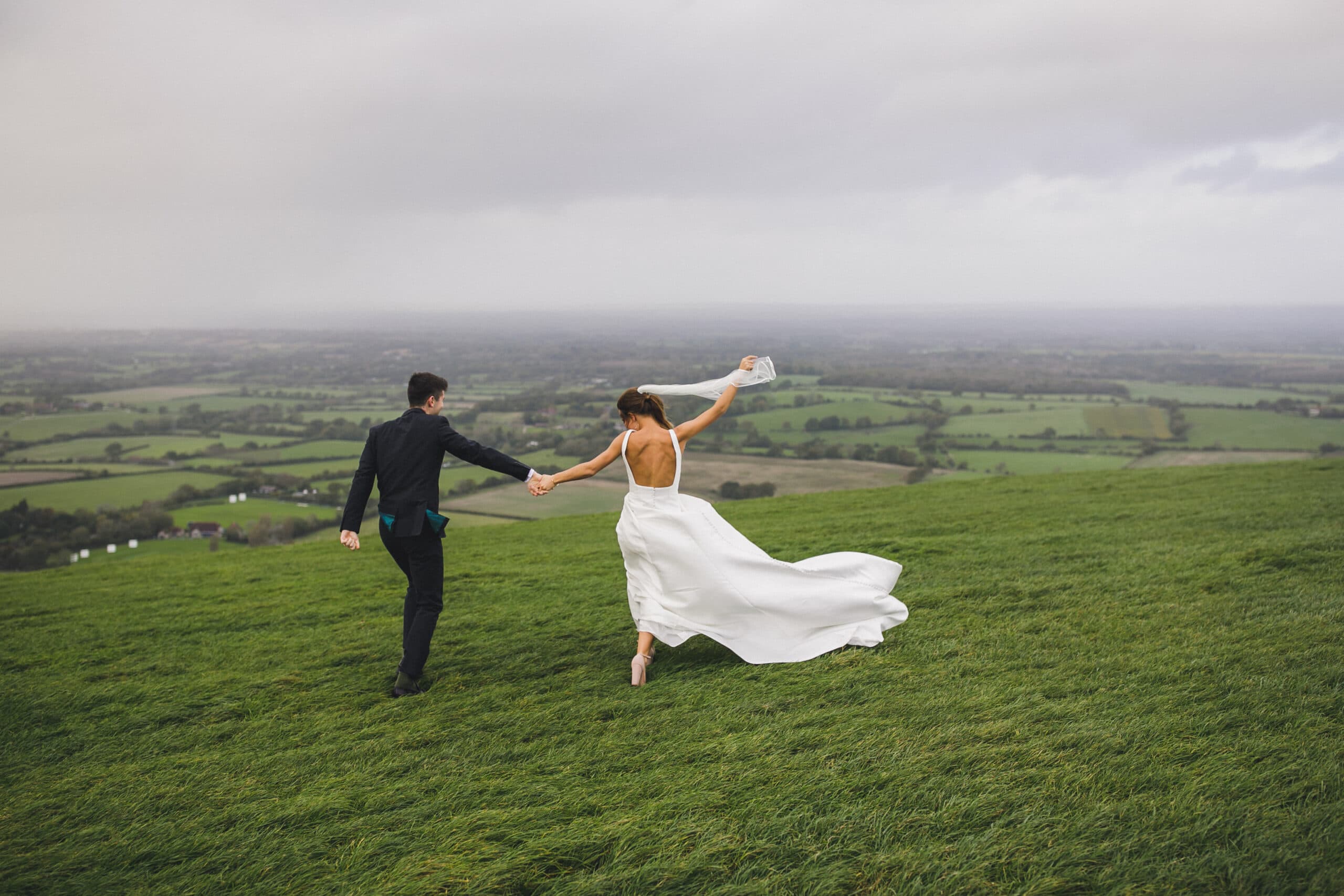 A bride and groom joyfully running on a grassy hill in Brighton